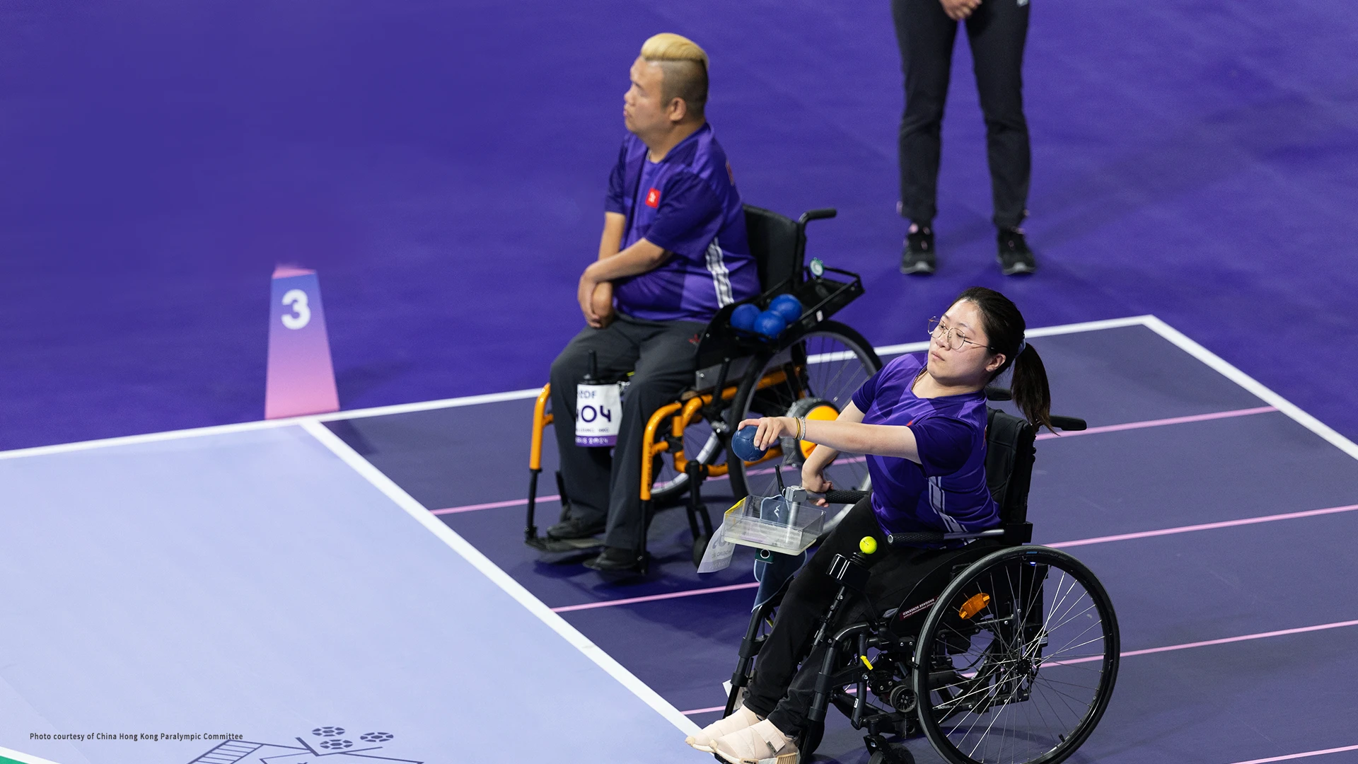 A man and a woman in wheelchairs, both in blue uniforms, with one of them about to throw a ball.
